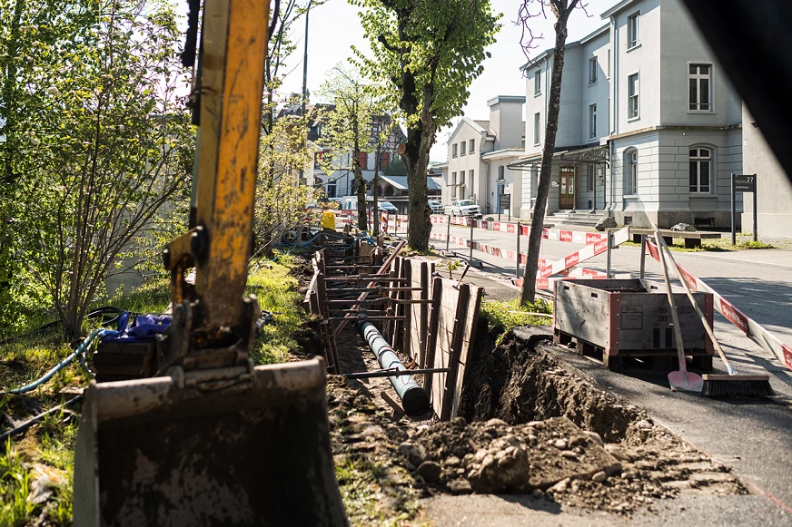 Leitungsarbeiten im Strassenbereich Baustelle entlang einer Strasse mit offenem Leitungsgraben, verlegten Rohren und einem Bagger vor mehreren Gebäuden.